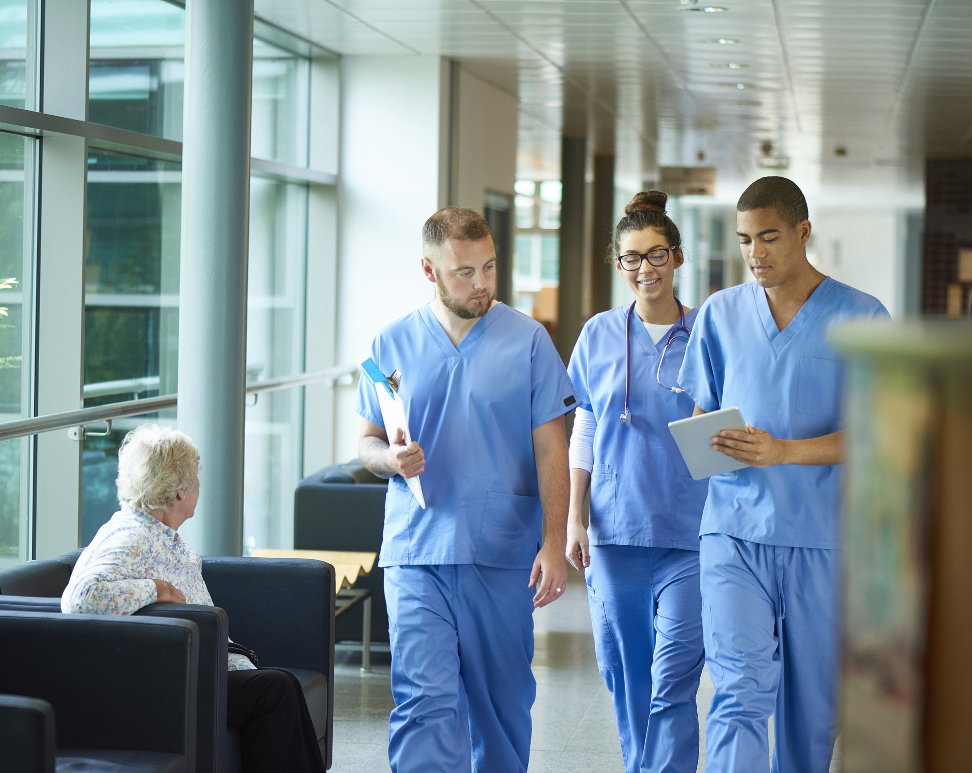three junior doctors walking along a hospital corridor discussing case and wearing scrubs. A patient or visitor is sitting in the corridor as they walk past.