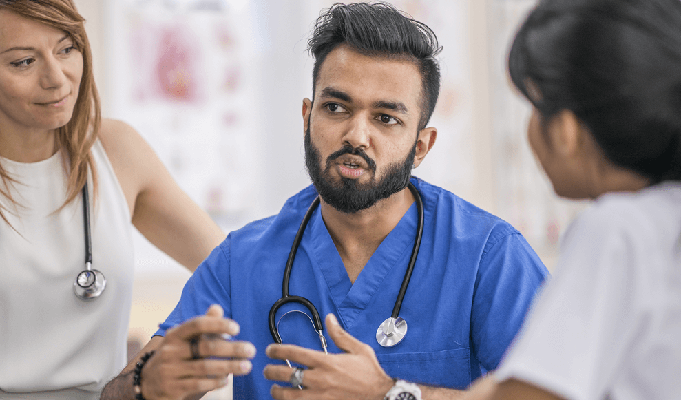 A group of three ethnically diverse medical professionals meet to discuss a patient file. They are seated together at a desk and each wearing their medical attire and stethoscopes. The focus is on a man if Indian decent who is leading the meeting.