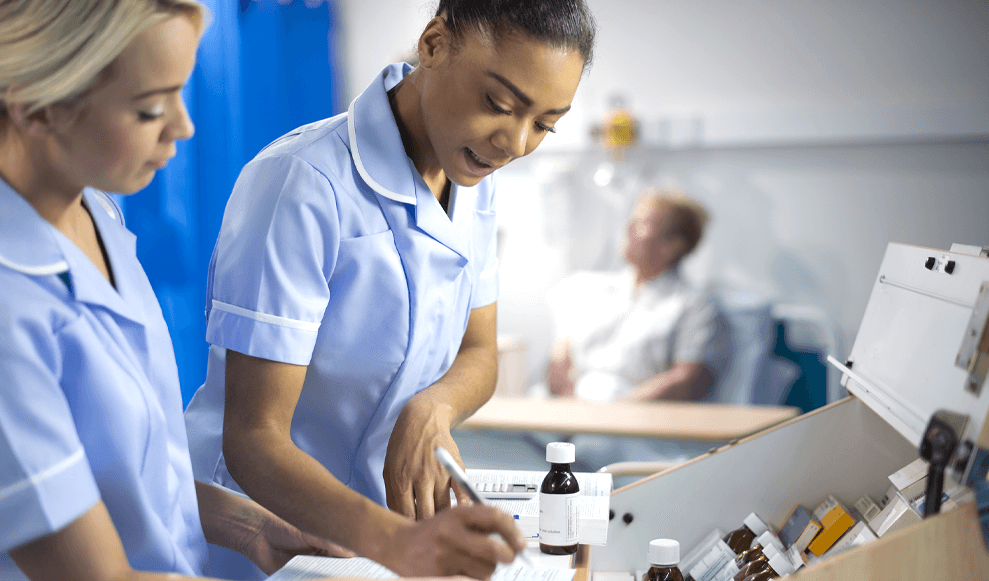 Two Nurses filling out paper work at a hospital ward dispensary, a patient is visible in the background waiting for medication