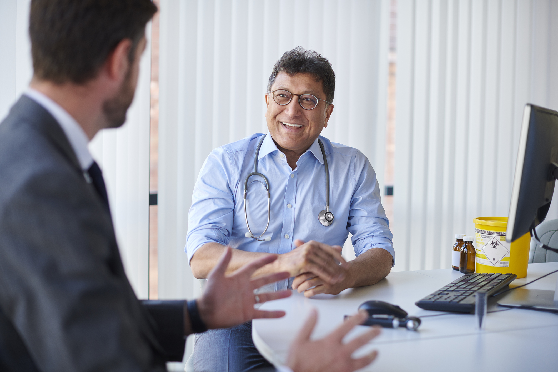 A GP is talking to a male patient in his office while sat at his desk