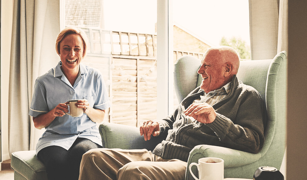 Senior man and his female carer having coffee and talking at home.