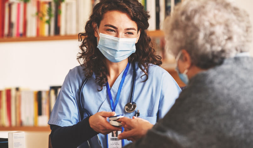 Doctor wearing surgical mask while visiting a patient at home. Senior woman sitting with doctor while doing coronavirus test and screening using oximeter. Rear view of old woman with grey hair giving finger to doctor for oximeter analysis.