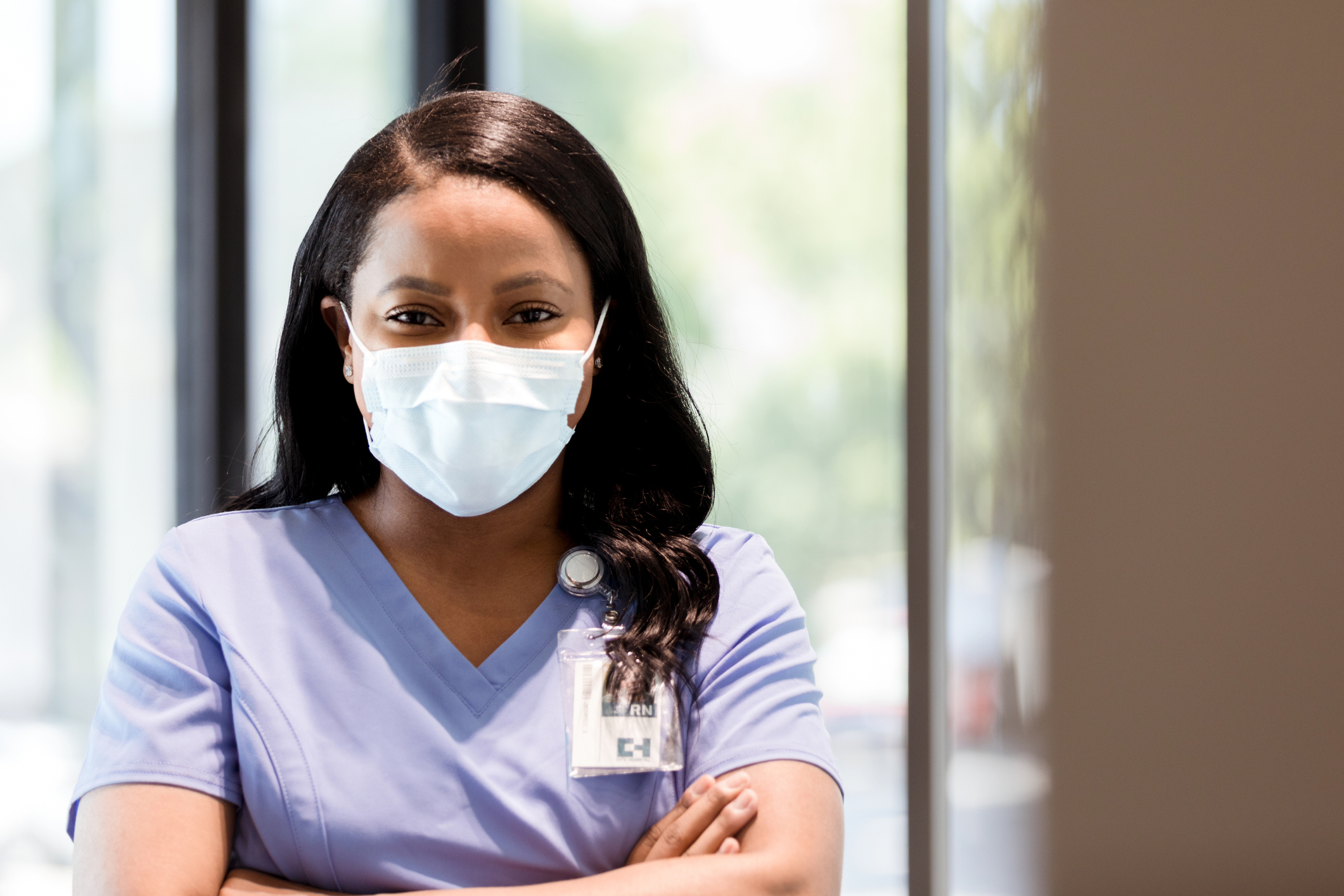 Portrait of a female nurse with her arms crossed, standing in front of a doctors office window smiling