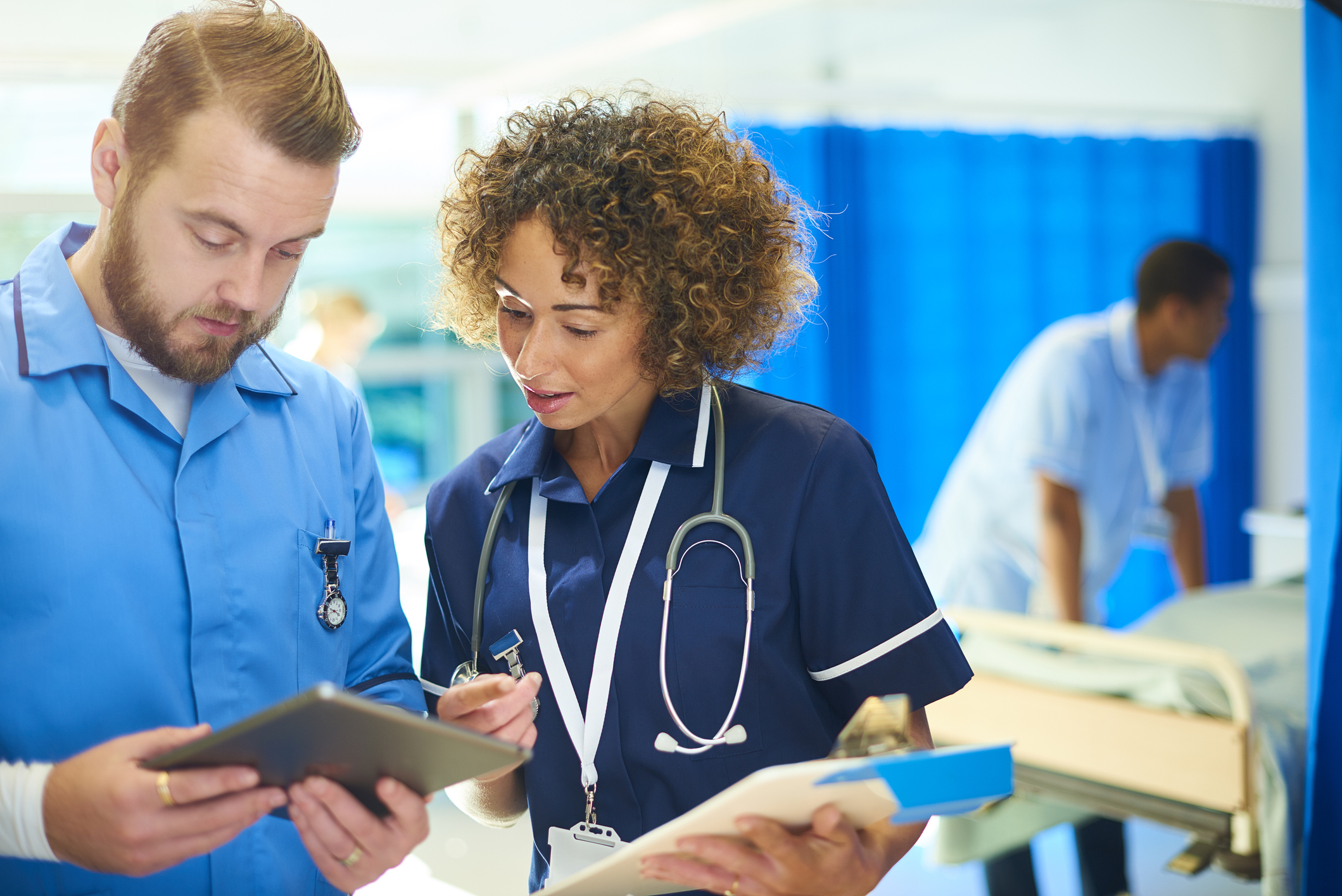 a nursing sister stands with a male nurse chatting about the medical chart that she is holding and cross referencing it with the digital tablet that he is holding. In the background a young nurse checks the dressing pack that she has got out of the trolley whilst a male nurse attends to a patient in bed .They are all wearing nurse uniforms consistent with the uk and the NHS