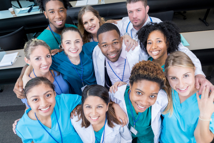 This is a view from above of a group of medical students looking up and smiling for the camera. They are standing in a lecture hall with table behind them.