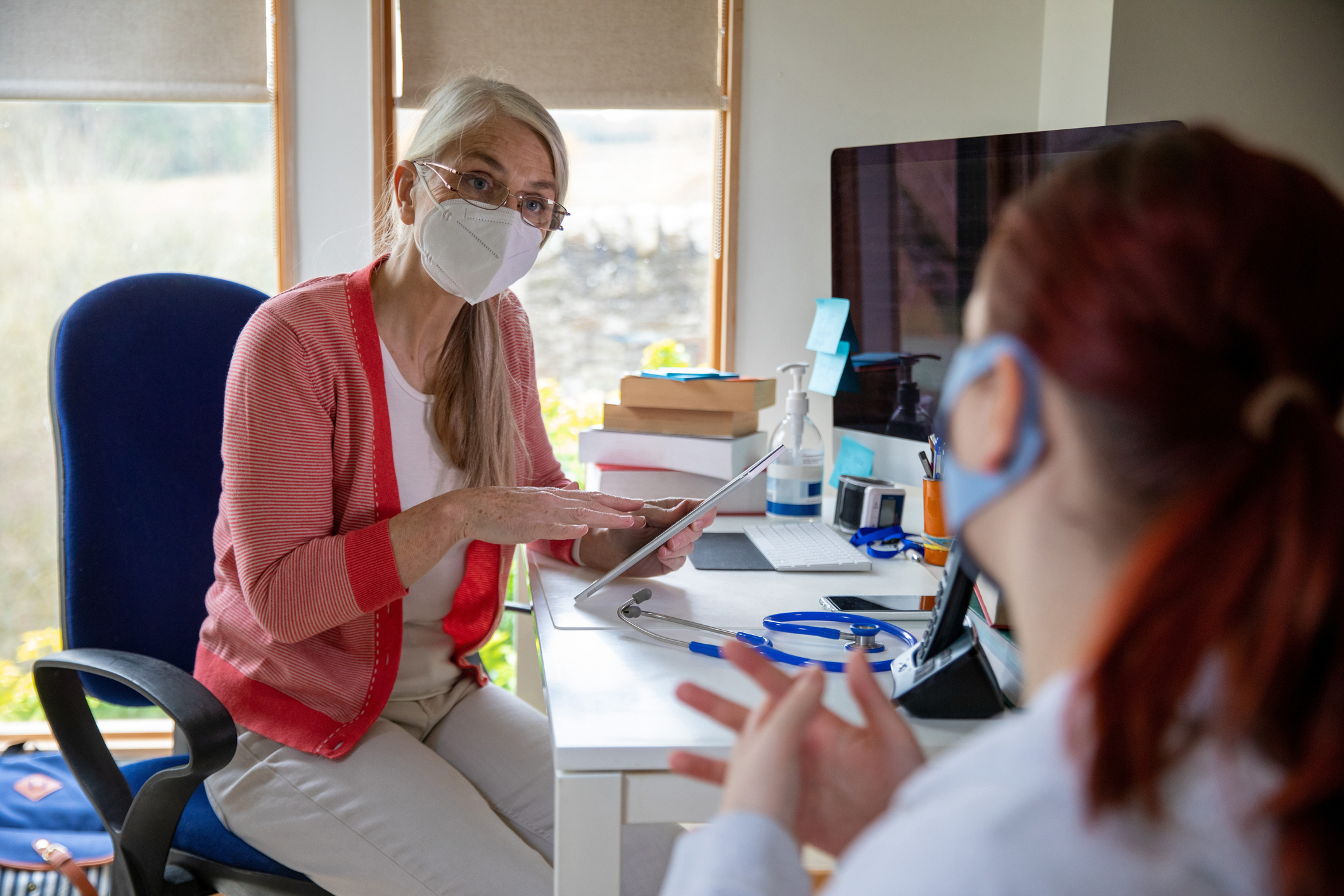 Doctor wearing a protective face mask working in a surgery during the Covid 19 pandemic. She is sitting at her desk with her medical tools in front of her while she holds a digital tablet. A patient is sitting with her, also wearing a face mask.