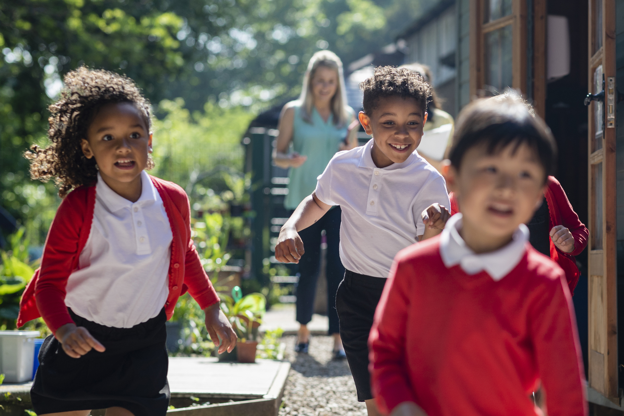 A front view of a group of school children including siblings running through the schoolyard in their uniform being followed by their teachers who are there to supervise and make sure they play safely outdoors.