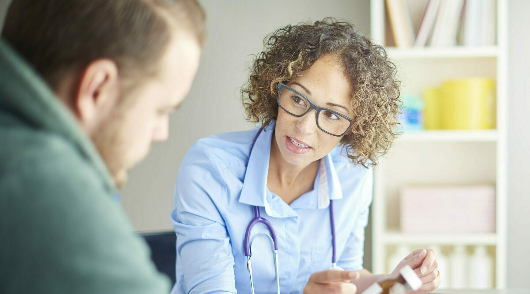 A female doctor sits at her desk and chats to a male patient about his current medication . She is dressed in a shirt with rolled up sleeves . They are both looking down at the pill bottle as she assesses his current dosage.