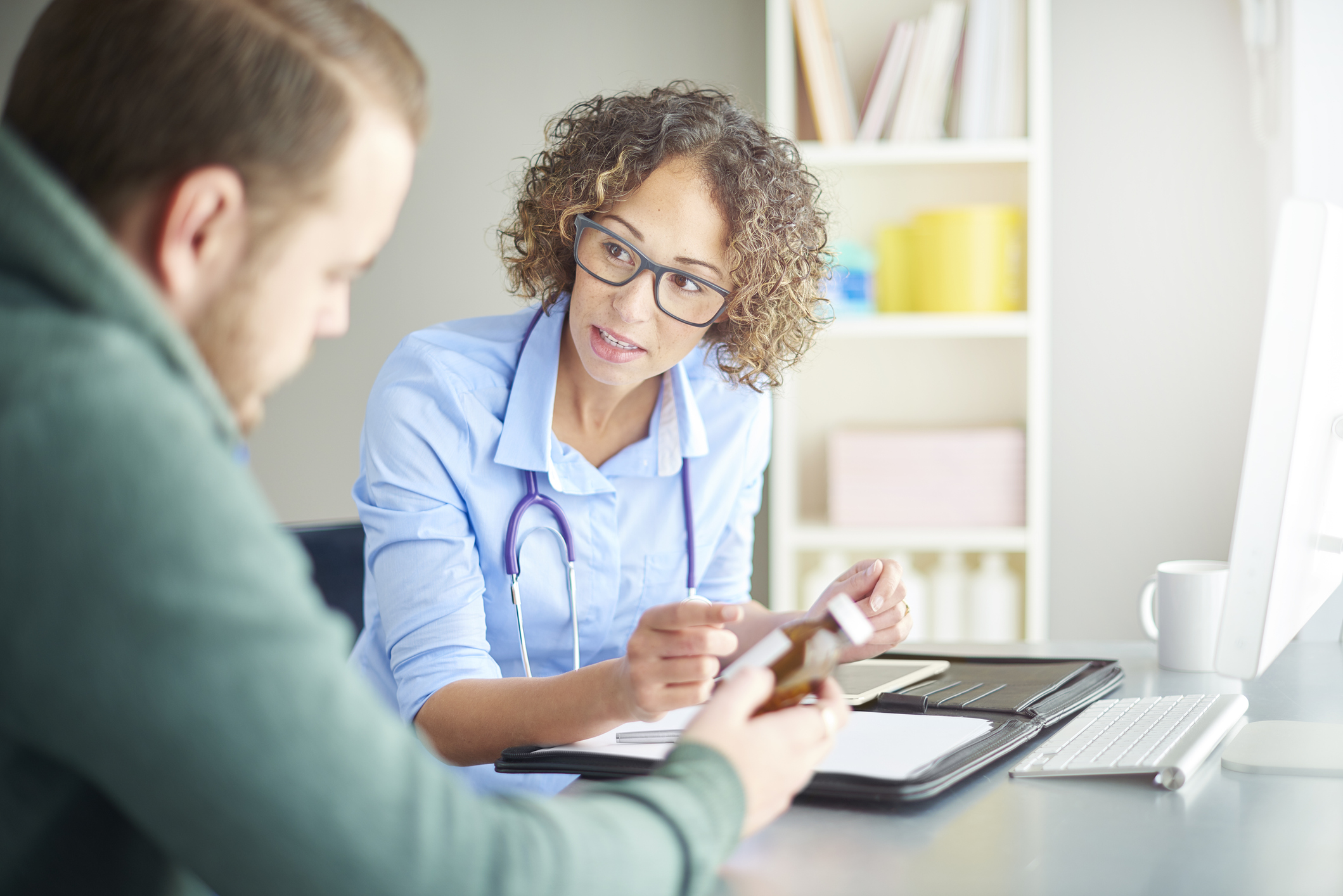 A female doctor sits at her desk and chats to a male patient about his current medication . She is dressed in a shirt with rolled up sleeves . They are both looking down at the pill bottle as she assesses his current dosage.