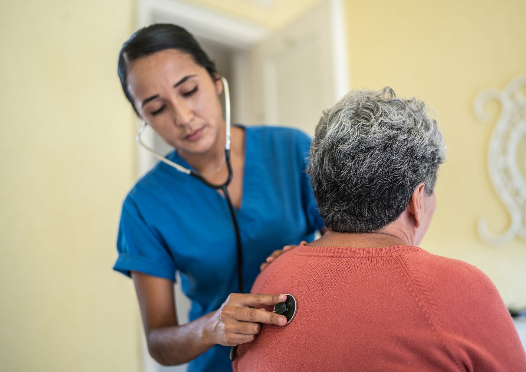 Nurse examining senior woman's lungs using stethoscope in the bedroom of a nursing home