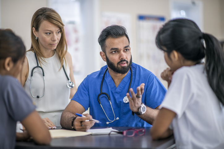 A group of doctors and nurses are in a meeting together. They are in a board room at the hospital. A male doctor is talking and his colleagues are listening to him.