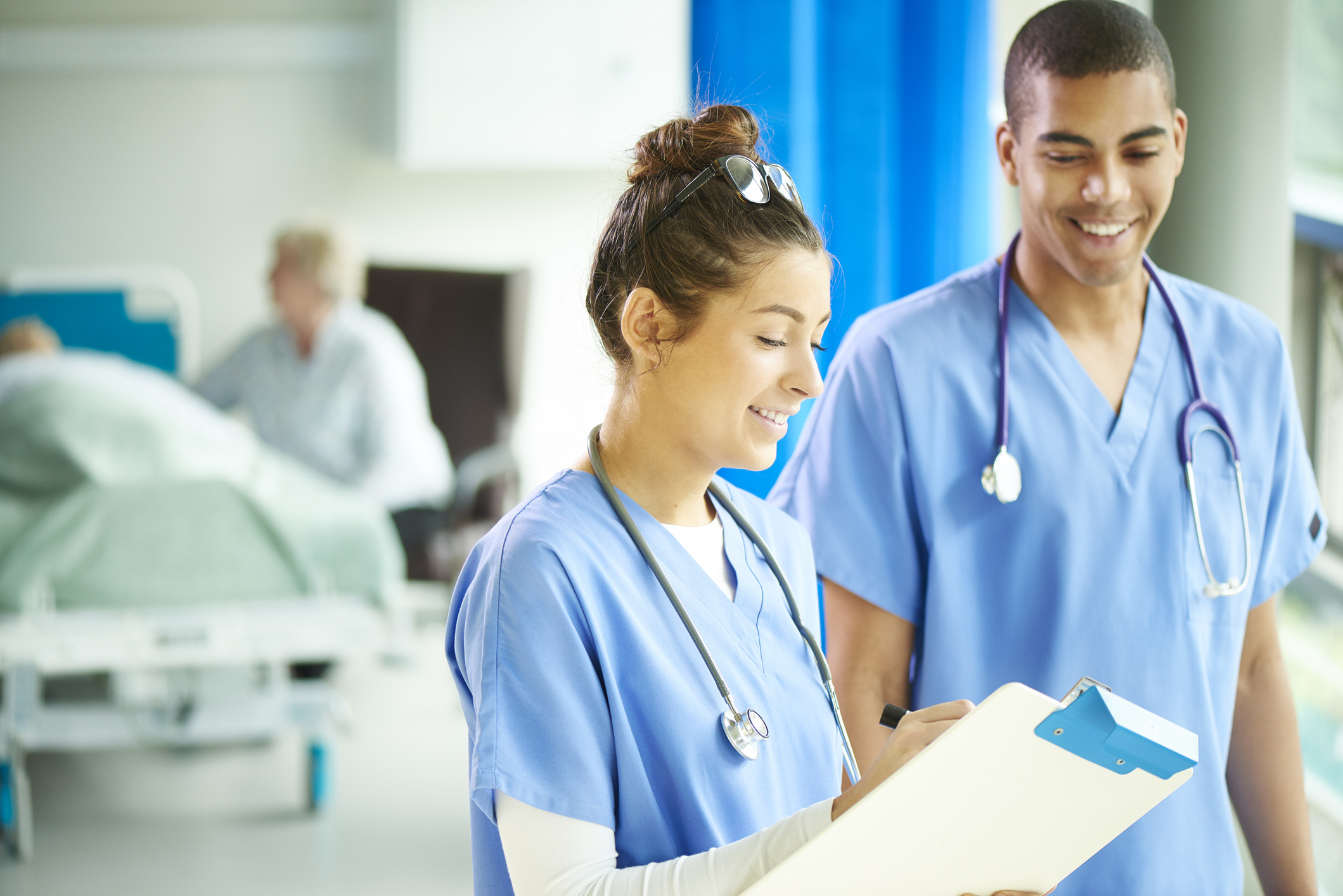 A young male and female nurse stand proudly on the ward and look to camera . In the background a patient is being attended by a nurse and their relative.