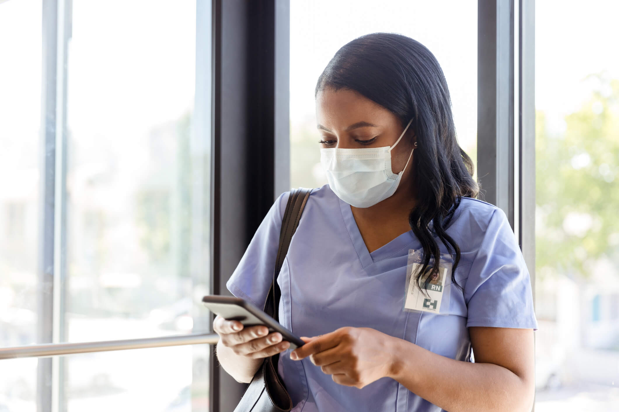 As a female nurse walks in a hospital hallway, she uses a smartphone to check her messages. She is wearing a surgical mask as she walks in the hospital.
