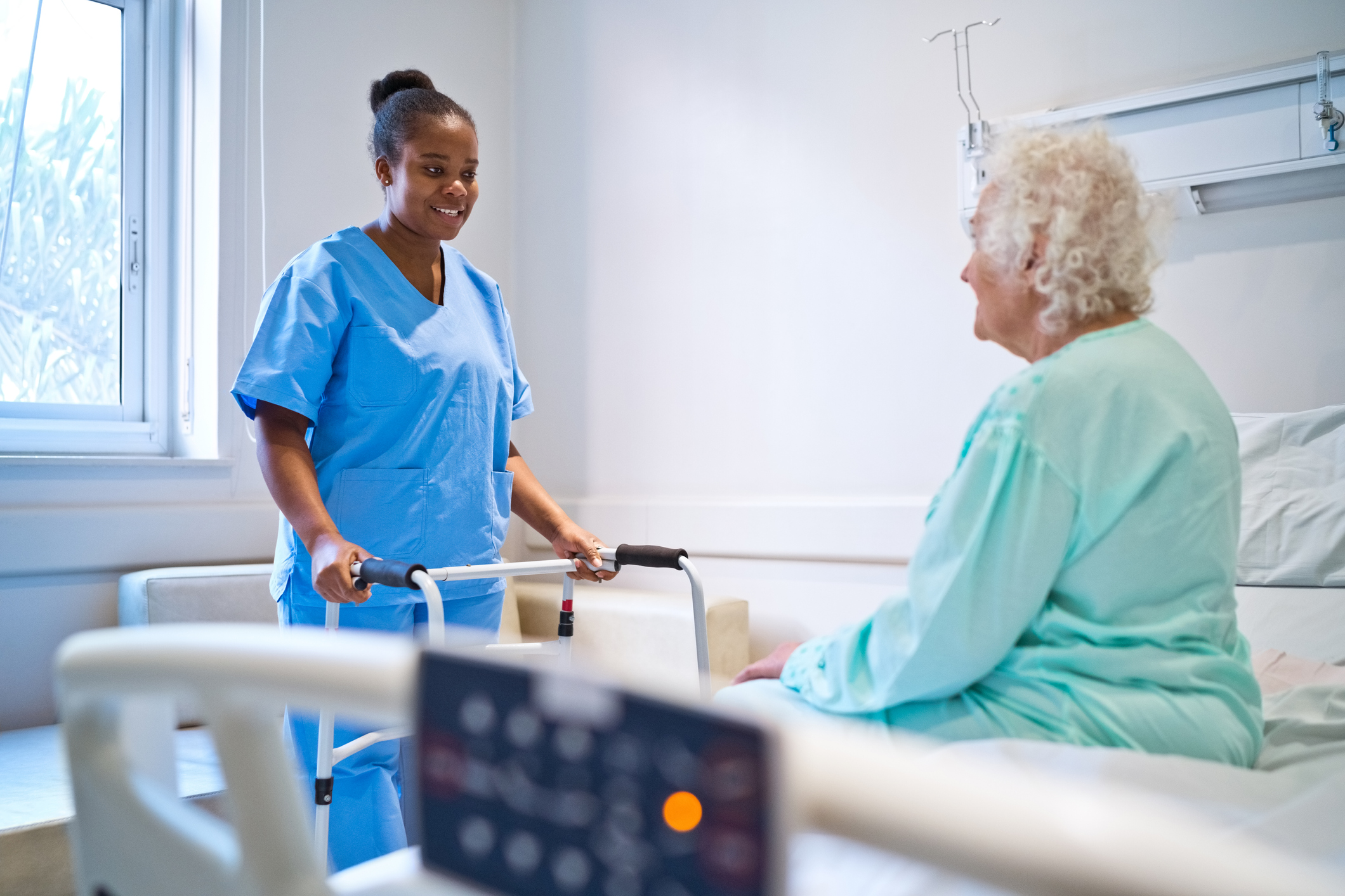 Smiling female nurse talking with patient sitting on bed in hospital ward