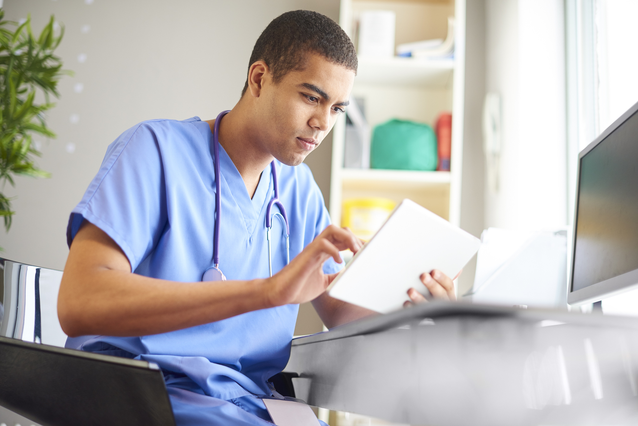 male gp sits at his desk looking at his digital tablet reading through patient notes, emails and looking through patients x-ray's. He is wearing blue scrubs and has his stethoscope around his neck.