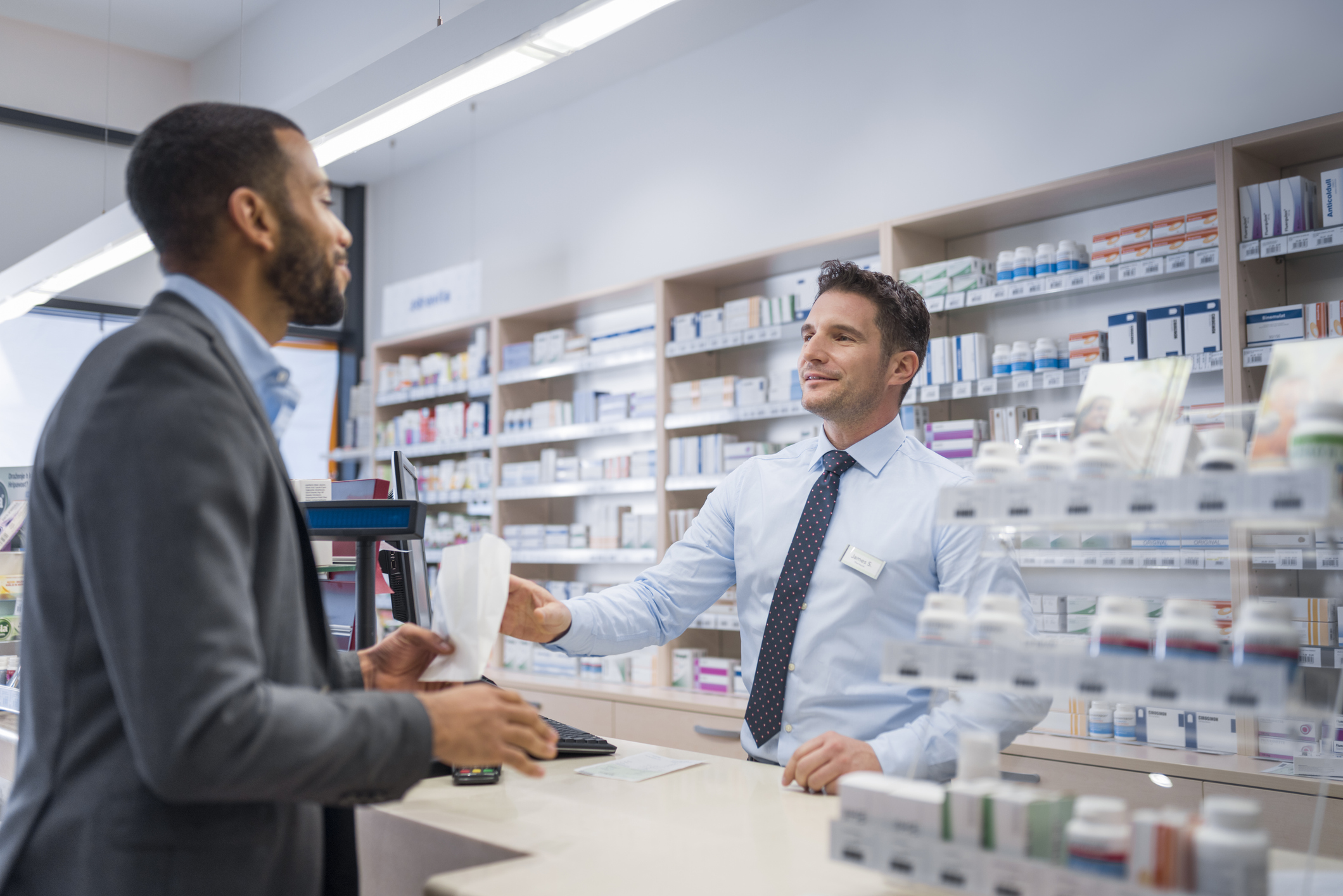 Male pharmacist giving medicine package to customer at counter in drugstore.