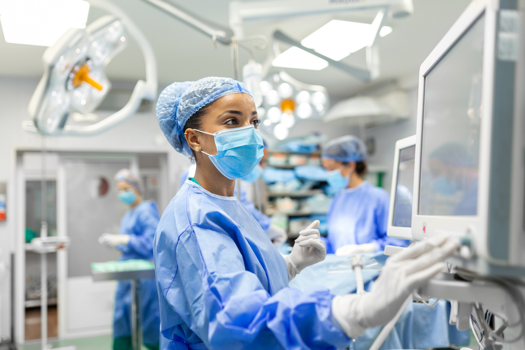 Anaesthetist Working In Operating Theatre Wearing Protectve Gear checking monitors while sedating patient before surgical procedure in hospital