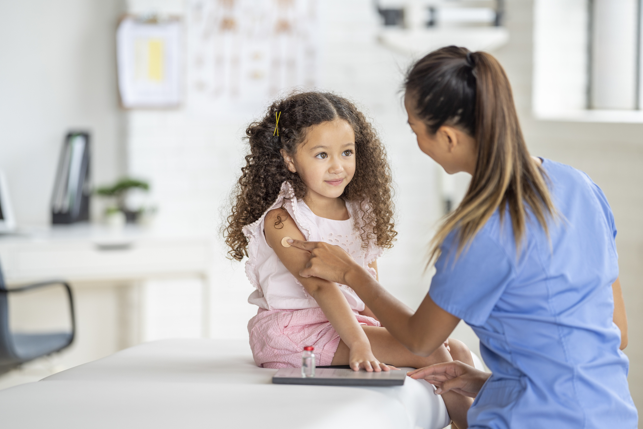 A sweet little girl of Latin decent, sits up straight on an exam table as her nurse places a bandage on her arm after giving her a vaccination. The nurse is wearing blue scrubs and smiling at the young girl to put her at ease.