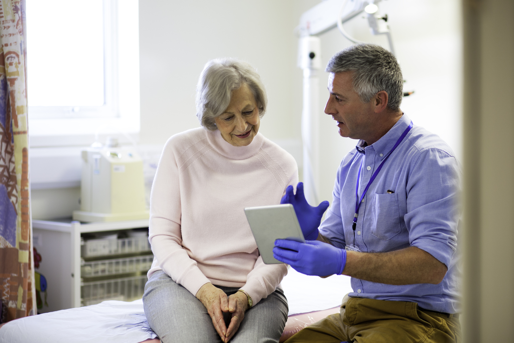 A male doctor sits beside an elderly female. They are in an examination room talking as he holds a digital tablet in one hand. The male doctor looks to be explaining something to his patient.