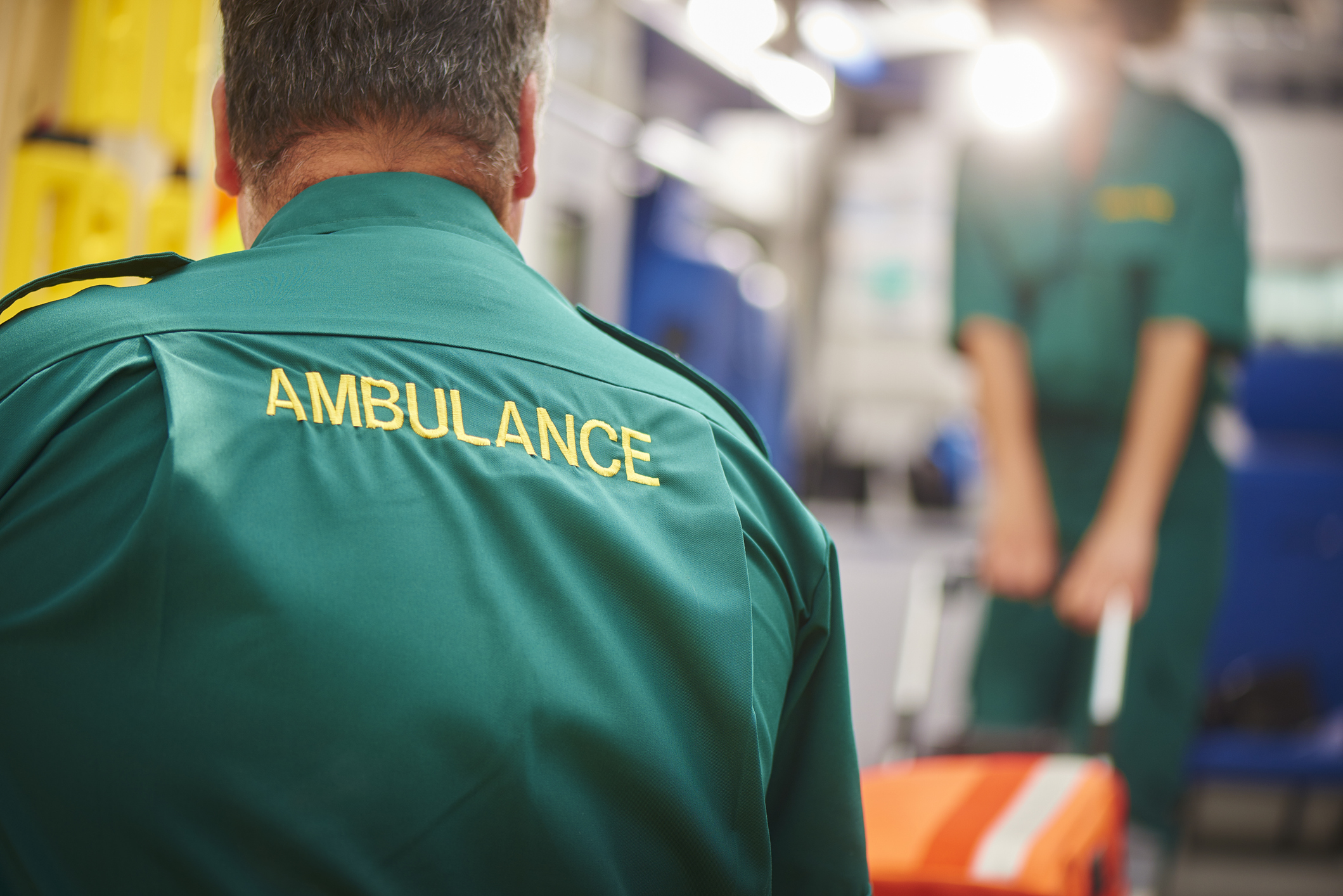 Two uk ambulance staff members pull a stretcher on or off of their ambulance . We are looking over the shoulder of the male paramedic focussing on the word ambulance on his back.They are wearing a green ambulance uniform typical of uk paramedics. The female paramedic and the back of the ambulance are defocussed.