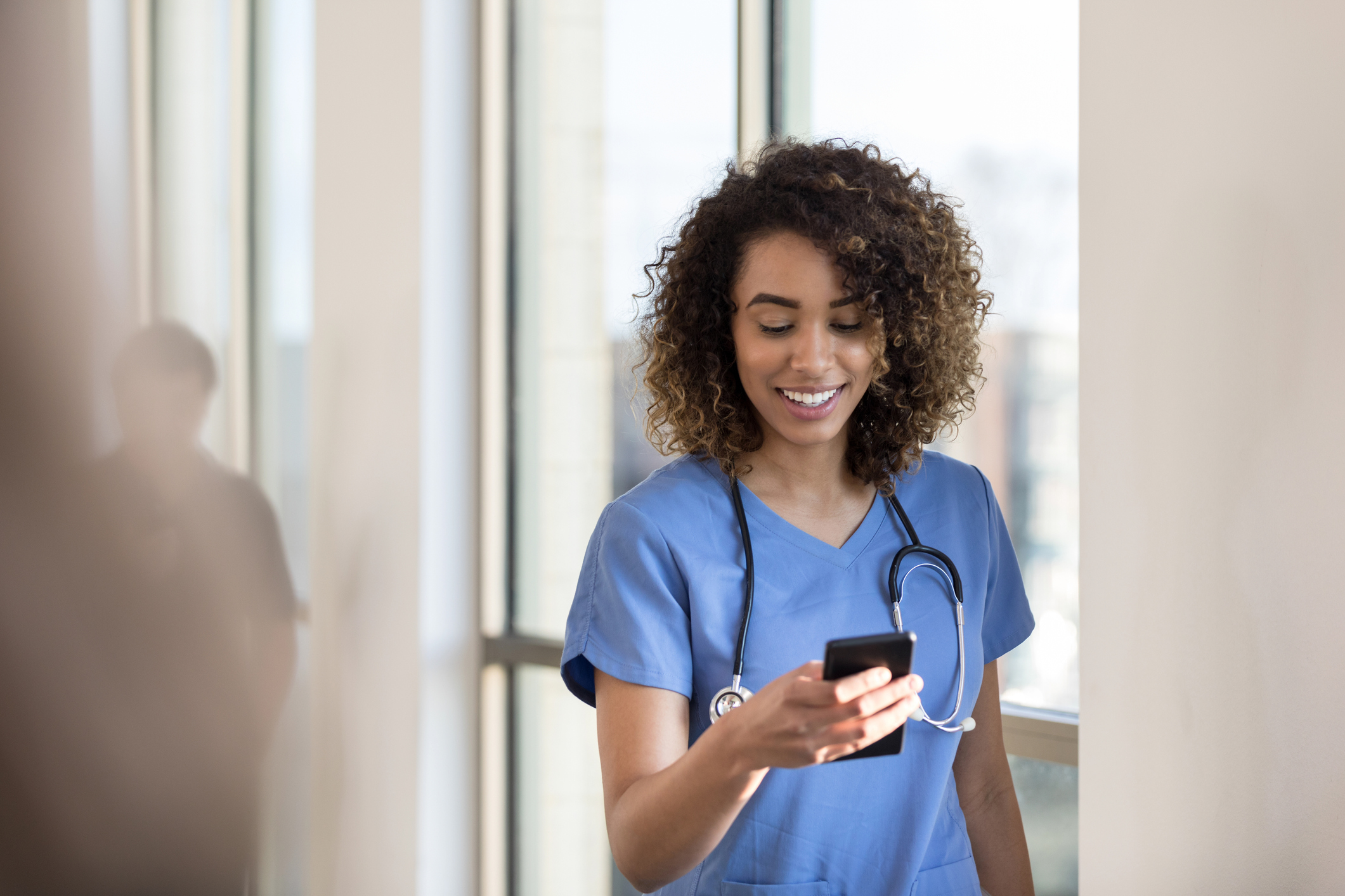 Cheerful Nurse checks messages on smartphone while in hospital hallway