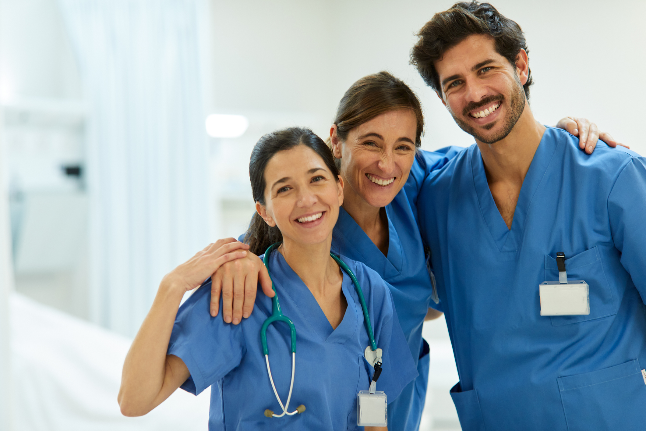 A trio of happy healthcare workers are grouped together posing for a photograph, they are on a hospital ward and wearing their blue scrubs