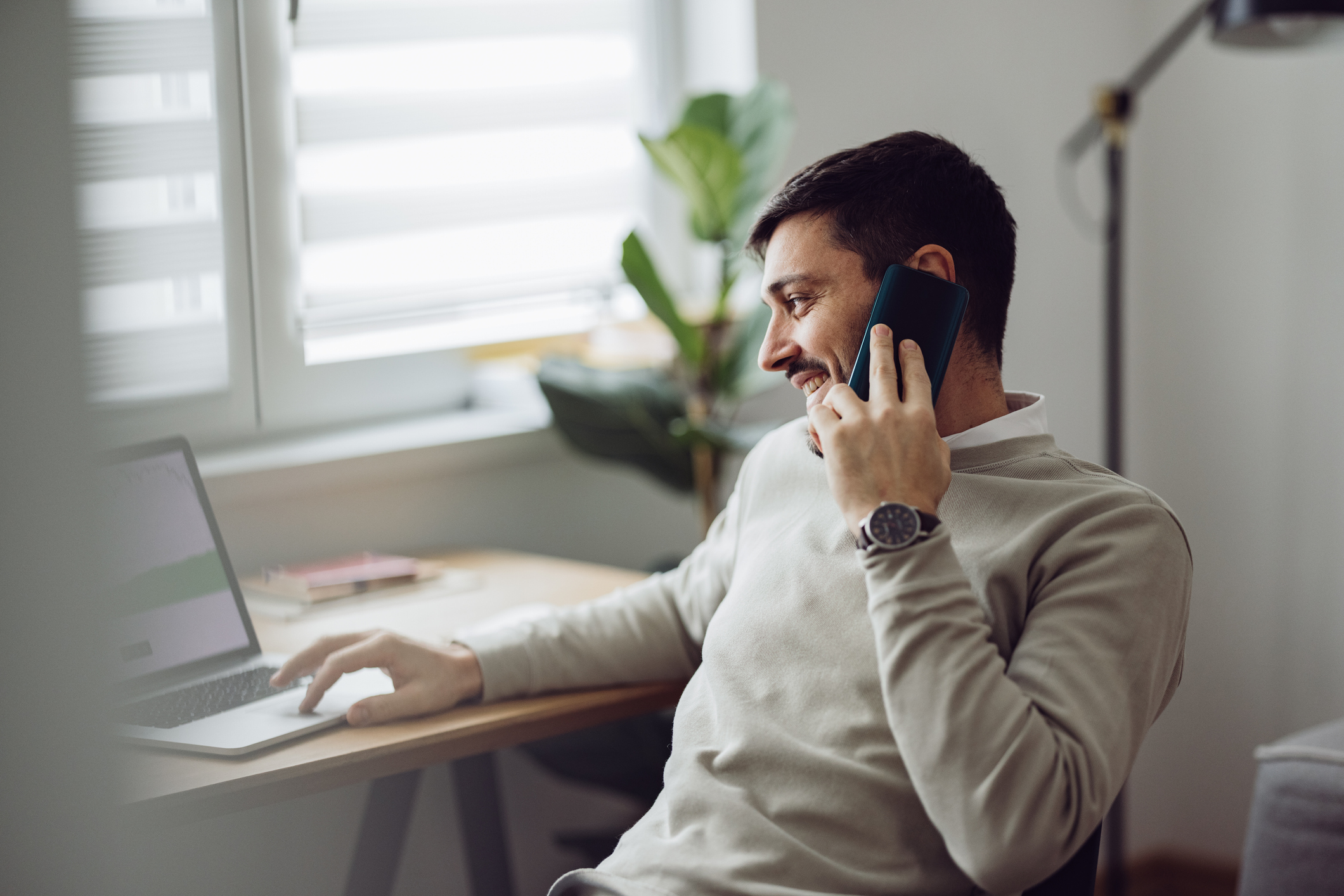 Caucasian businessman is talking on his smartphone while using a laptop for online trading and following the stock market from home. It's morning or later in the day.