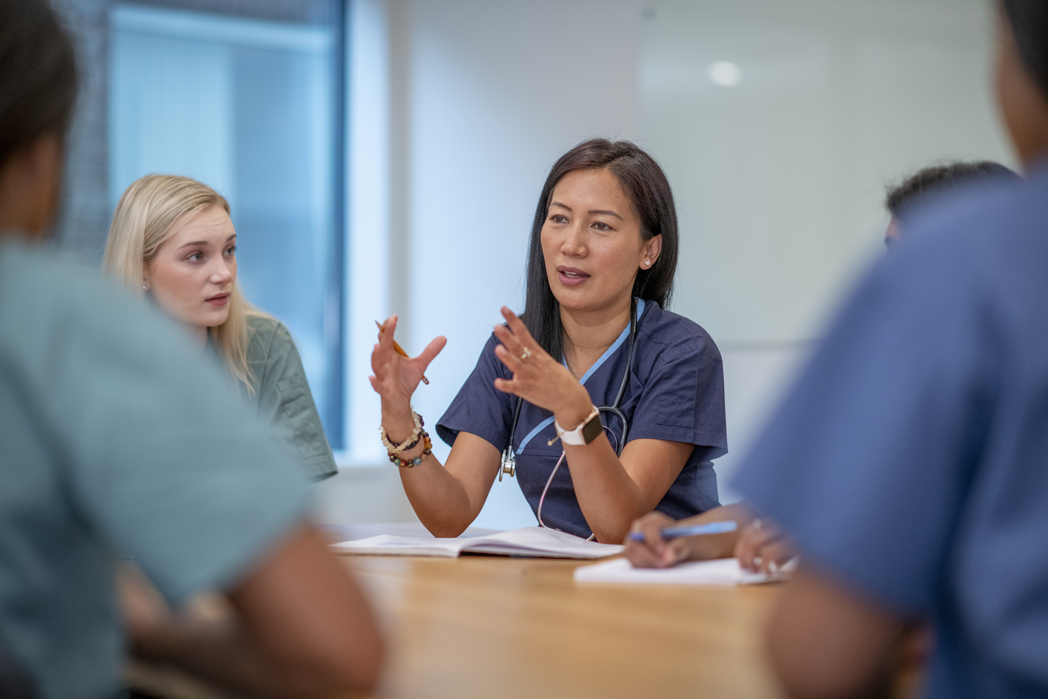 A small group of Nurse sit at a boardroom table as they meet to discuss patient cases. They are each dressed professionally in scrubs and have stethoscopes around their necks as they discuss plans of care for each case.