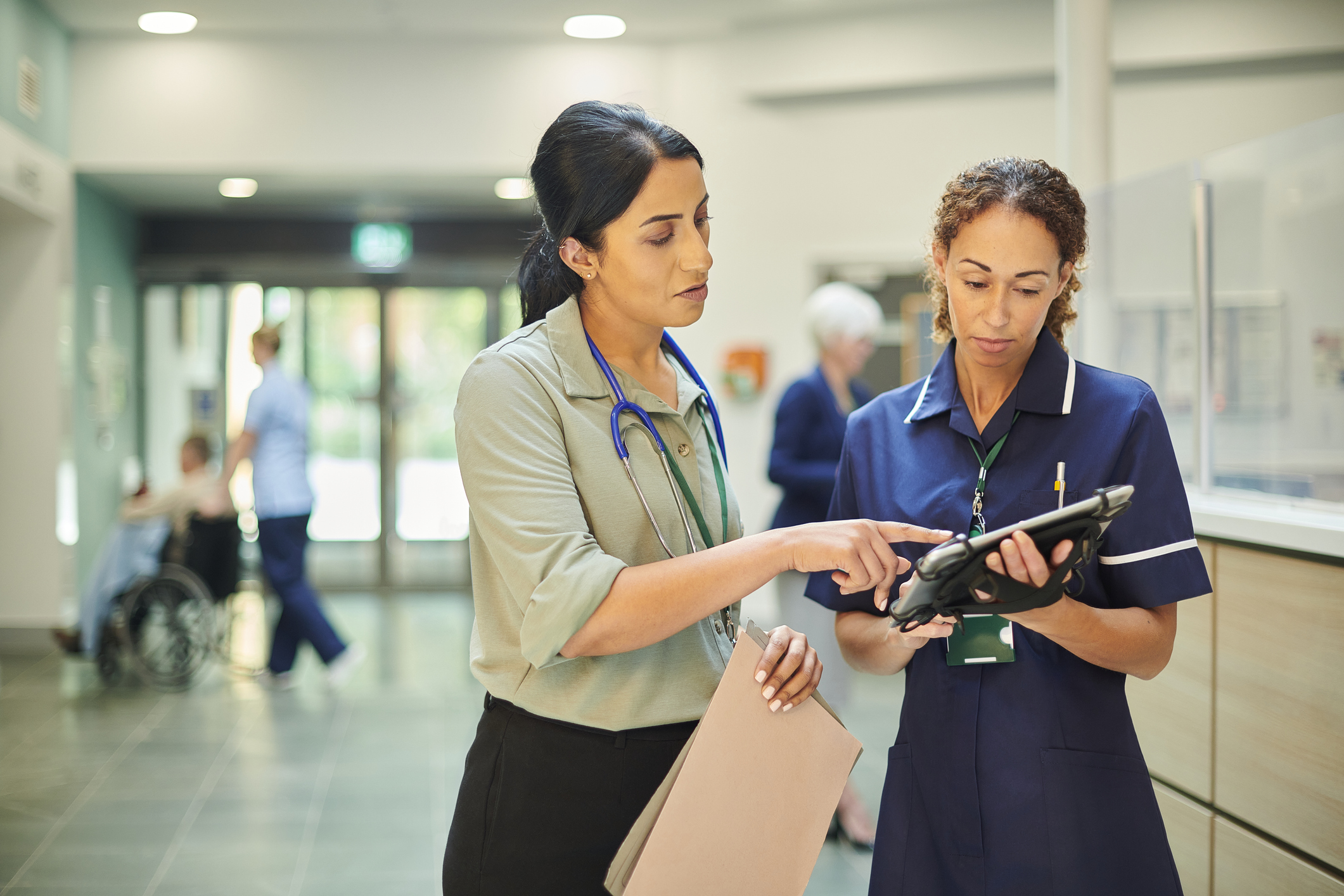 A hospital nurse is discussing a case with a consultant in a hospital lobby, they are looking at a digital tablet device