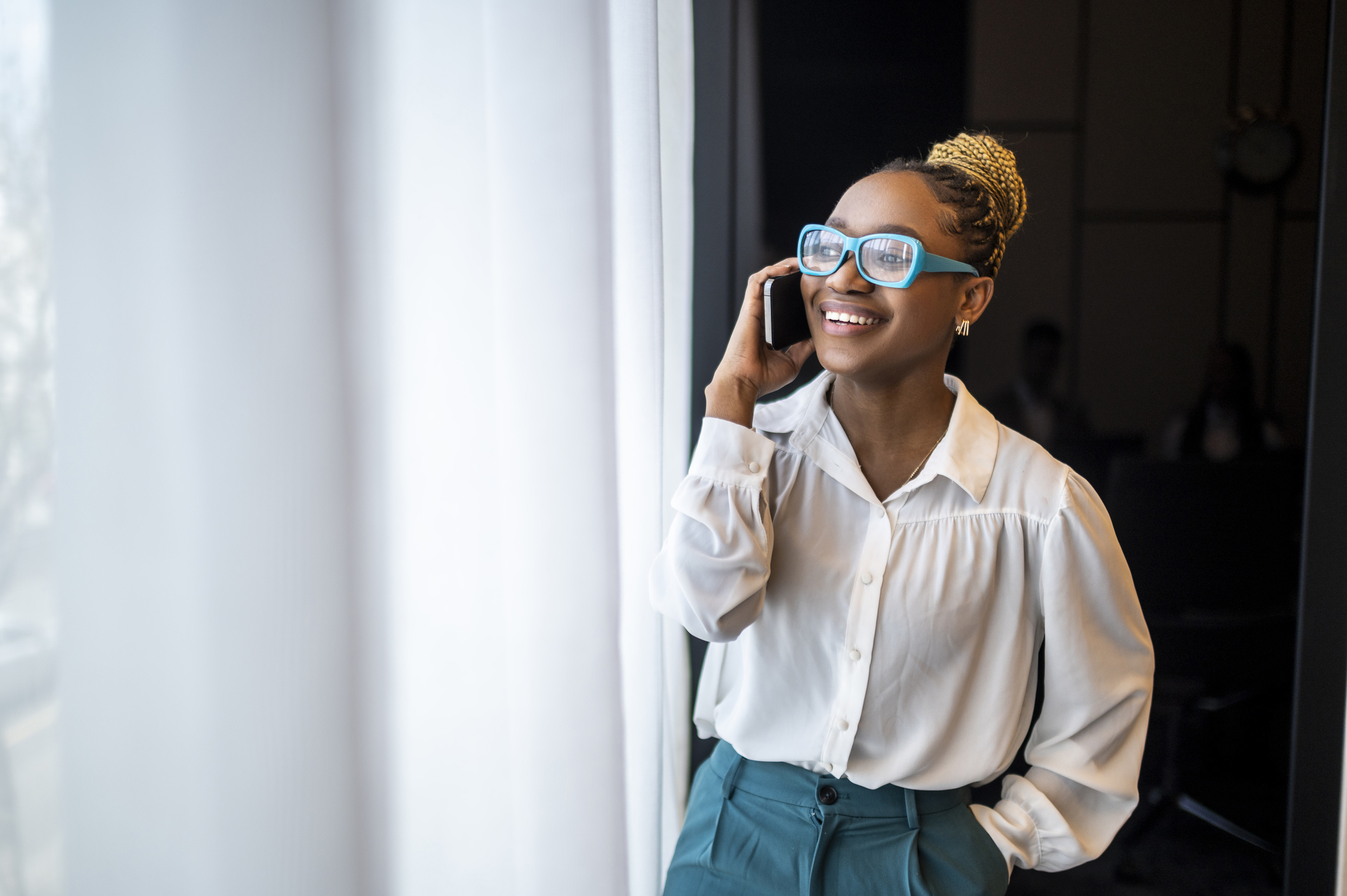 A beautiful black woman with blue eyeglasses and a bun in white shirt and dark green pants seen standing by the window and smiling while talking on the phone during a break between meetings.