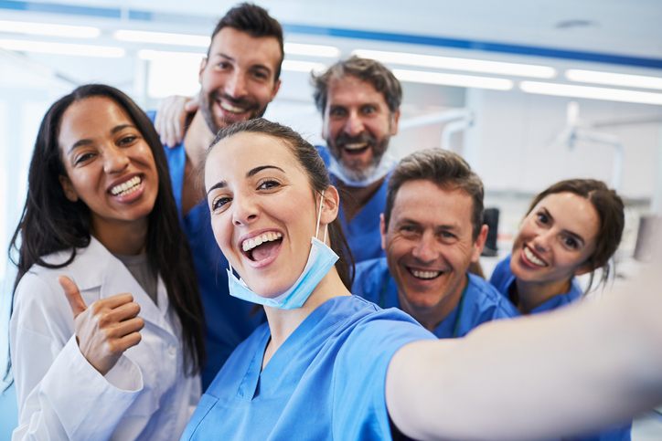 A group of healthcare workers are grouped together taking a selfie in their work scrubs