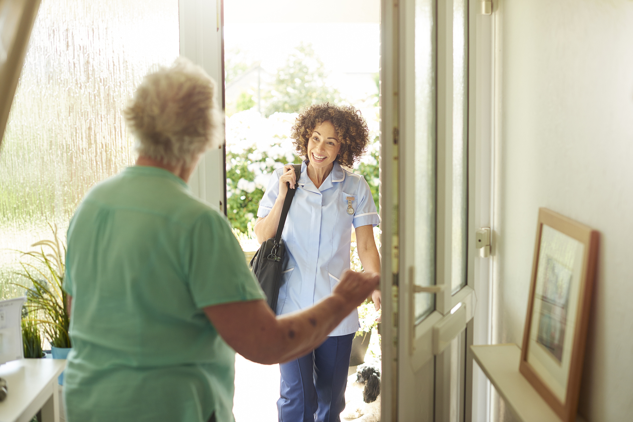 a care worker or medical professional makes a house call to a senior patient at her home . They are standing at the front door as she enters.