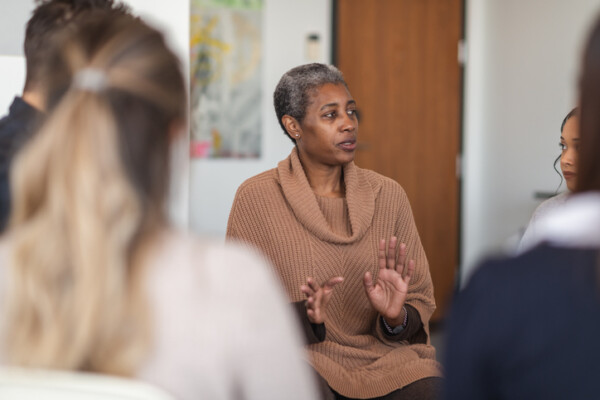 A black mature adult woman is a psychologist. The mental health professional is leading a group therapy session. The multi-ethnic group of adults are sitting in a circle in a clinic. The therapist is speaking and the attendees are listening to her advice.