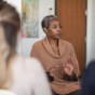 A black mature adult woman is a psychologist. The mental health professional is leading a group therapy session. The multi-ethnic group of adults are sitting in a circle in a clinic. The therapist is speaking and the attendees are listening to her advice.